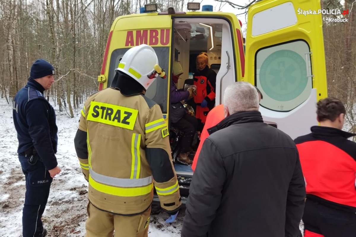 Na zdjęciu widać osobę bezdomną w karetce pogotowia, obok stoją strażacy, umundurowany policjant i mężczyzna w cywilnym ubraniu