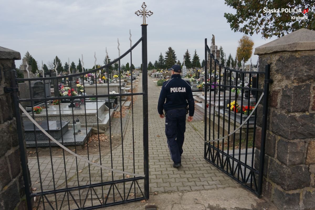 Na pierwszym planie widzimy otwarte prawe skrzydło bramy cmentarza. Dalej policjanta, który idzie ścieżką cmentarza pomiędzy grobami.