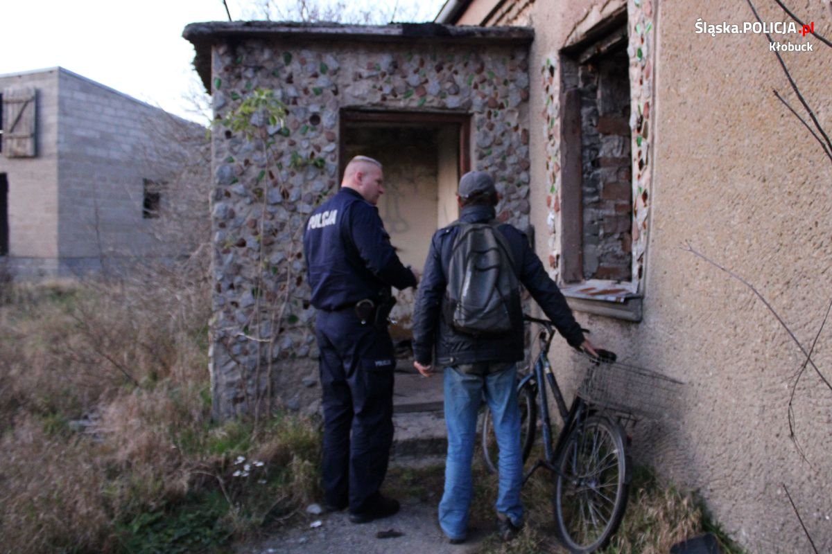 Na zdjęciu widzimy fragment budynku, przed którego wejściem stoi policjant i rozmawia z mężczyzną stojącym obok niego. Mężczyzna trzyma dłoń na rowerze.