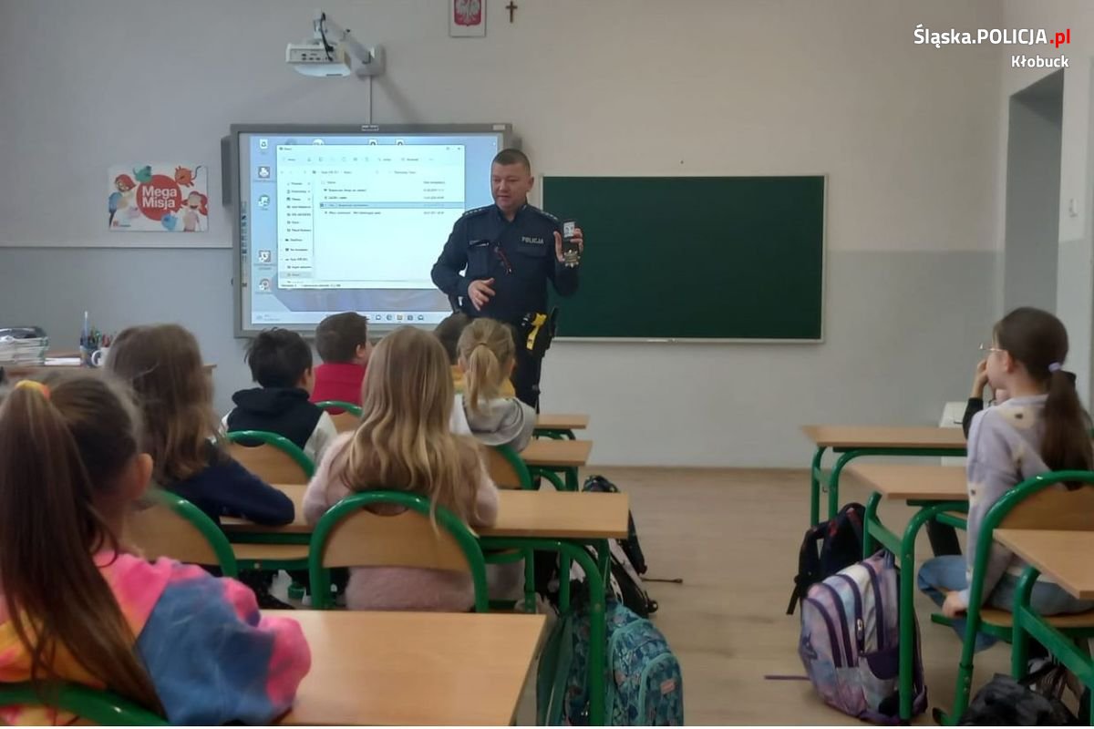 Zdjęcie przedstawia policjanta, który prezentuje siedzącym przed nim dzieciom jak wygląda legitymacja służbowa policjanta.