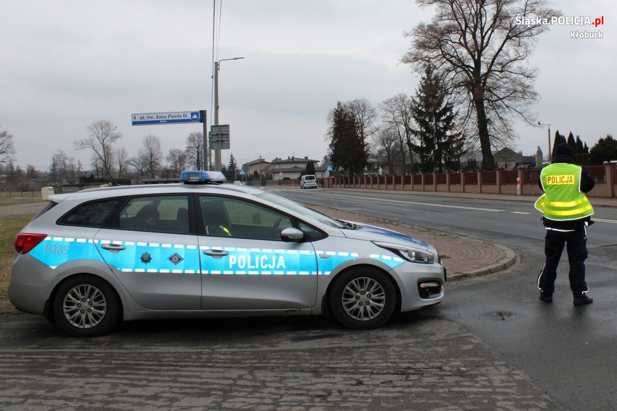 Zdjęcie przedstawia umundurowanego policjanta mierzącego prędkość przejeżdżających pojazdów oraz oznakowany radiowóz.