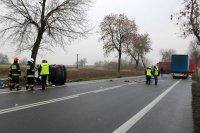 po lewej stronie fotografii widać policjanta oraz strażaków, którzy stoją obok rozbitego osobowego forda; po prawej stronie widać policjantów wykonujących czynności na miejscu zdarzenia a w tle dwa pojazdy ciężarowe