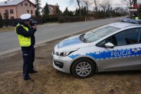 po lewej stronie stoi policjant, który urządzenie mierzy prędkość nadjeżdżającego pojazdu, po prawej stronie znajduje się radiowóz a za nim drugi policjant