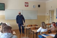 umundurowany policjant stoi w klasie przodem do uczniów. którzy siedzą w ławkach