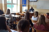 umundurowany policjant stoi w klasie siedzi za biurkiem przodem do uczniów. którzy siedzą w ławkach