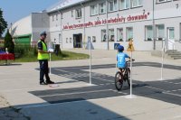 na zdjęciu widać chłopaka, który jedzie na rowerze po torze mobilnego miasteczka ruchu drogowego, obok po lewej stronie stoi umundurowany policjant