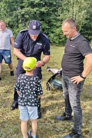 policjant zakłada opaskę na rękę dziecka, które ma na głowie żółtą czapkę z daszkiem, obok stoi mężczyzna