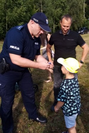 policjant zakłada opaskę na rękę dziecka, które ma na głowie żółtą czapkę z daszkiem, obok stoi mężczyzna