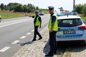 dwóch umundurowanych policjantów stoi przy radiowozie, jeden daje znak tarczą do zatrzymywania pojazdów