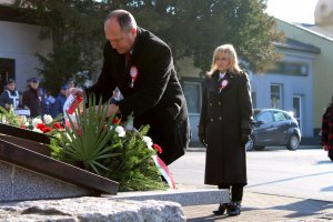 mężczyzna składa biało czerwone kwiaty przy pomniku w tle kobieta w płaszczu