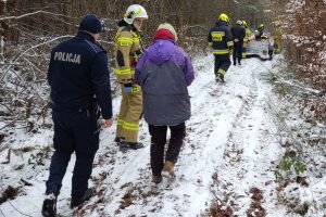 bezdomna kobieta idzie drogą leśną, obok umundurowany policjant i strażak