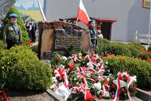 wieńce biało czerwonych kwiatów przed pomnikiem. obok stoją harcerze