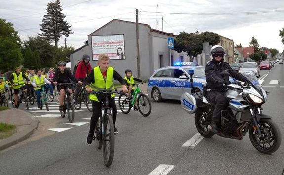 rowerzyści w kamizelkach odblaskowych przejeżdżają przez jezdnię, w tle radiowóz, obok policyjny motocykl
