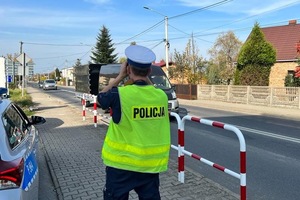 Policjant ruchu drogowego obserwuje przez lornetkę kierowców w przejeżdżających pojazdach. Po jego lewej stronie widać fragment radiowozu.