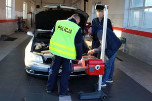 Wewnątrz stacji diagnostycznej policjant i diagnosta sprawdzają stan i ustawienie świateł w pojeździe za pomocą specjalistycznego urządzenia. Obok stoi kierowca kontrolowanego pojazdu.