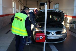 Wewnątrz stacji diagnostycznej policjant i diagnosta sprawdzają stan i ustawienie świateł w pojeździe za pomocą specjalistycznego urządzenia.