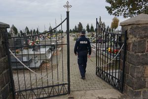 Na pierwszym planie widzimy otwarte prawe skrzydło bramy cmentarza. Dalej policjanta, który idzie ścieżką cmentarza pomiędzy grobami.