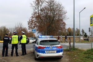 Po lewej stronie widzimy trzech policjantów. Po prawej stronie policyjny radiowóz z włączonymi światłami błyskowymi. W tle cmentarz.