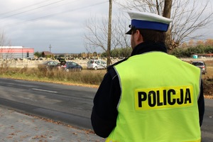 Widzimy korpus stojącego tyłem policjanta ruchu drogowego. W tle cmentarz.