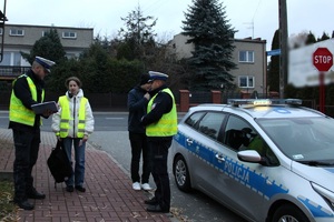 Zdjęcie przedstawia dwóch umundurowanych policjantów, pieszą w kamizelce odblaskowej oraz radiowóz.