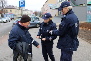 Zdjęcie przedstawia dwie umundurowane policjantki, które wręczają opaskę odblaskową pieszemu.