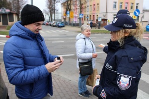Zdjęcie przedstawia dwie umundurowaną policjantkę, która wręcza opaskę odblaskową pieszemu.