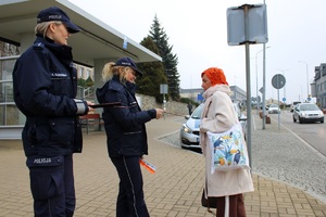 Zdjęcie przedstawia dwie umundurowane policjantki, które wręczają opaskę odblaskową pieszej.