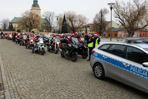 Zdjęcie przedstawia radiowóz, za którym stoją policjanci, motocykle i motocykliści w przebraniach Mikołaja.