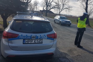 Zdjęcie przedstawia oznakowany policyjny radiowóz oraz umundurowanego policjanta, który mierzy prędkość przejeżdżającego samochodu.