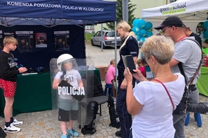 Zdjęcie przedstawia promocyjne stoisko policyjne, policjantkę oraz chłopca z tarczą policyjna i w hełmie.