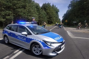 oznakowany radiowóz stoi przy drodze, którą jadą rowerzyści, obok stoi policjant w mundurze