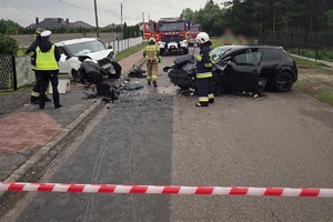 miejsce wypadku drogowego, widoczne dwa pojazdy z uszkodzeniami: fiat oraz seat, obok stoi umundurowany policjant oraz strażacy