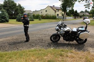 Policjant mierzy prędkość nadjeżdżającym pojazdom. Obok stoi policyjny motocykl.