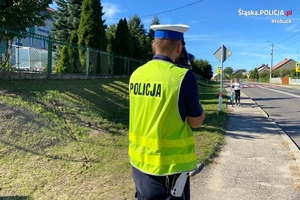 Policjant, dziewczynka prowadząca rower oraz przejście dla pieszych przed szkołą.