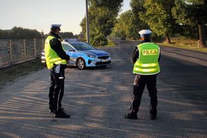 dwóch umundurowanych policjantów stoi przy radiowozie zaparkowanym na przydrożnym parkingu