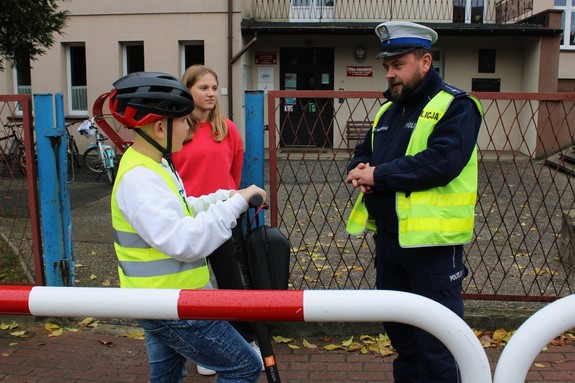 umundurowany policjant rozmawia przed szkołą z uczennicą i uczniem trzymającym hulajnogę