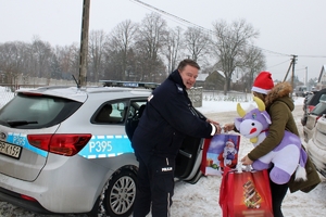 Zdjęcie przedstawia policjanta, który przekazuje paczki prezentowe kobiecie w czapce Mikołaja.