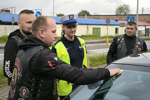 Zdjęcie przedstawia policjanta oraz motocyklistę rozmawiających z kierowcą samochodu i przklejających naklejkę na szybie.