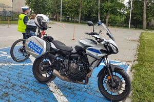 Zdjęcie przedstawia policyjny motocykl, policjanta oraz dziecko pokonujące na rowerze tor przeszkód.