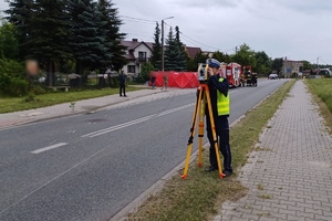 Umundurowany policjant prowadzi oględziny miejsca wypadku, stoi na drodze, w tle wóz strażacki oraz czerwony parawan strażacki