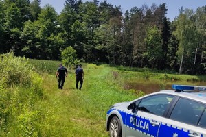 umundurowani policjanci sprawdzają zarośla przy zbiorniku wodnym, na zdjęciu widać również oznakowany radiowóz