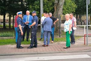 umundurowani policjanci wraz innymi osobami stoją na chodniku przy drodze