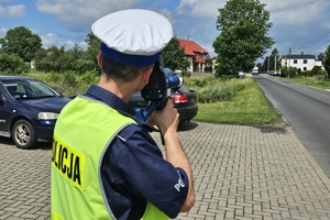 Zdjęcie przedstawia policjanta ruchu drogowego mierzącego prędkość nadjeżdżającego pojazdu.