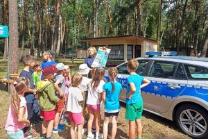 Zdjęcie przedstawia policjantkę pokazującą plakat dzieciom stojącym obok radiowozu.