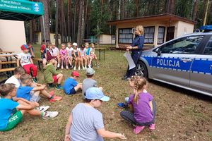 Zdjęcie przedstawia policjantkę stojąca obok radiowozu oraz siedzące dzieci.