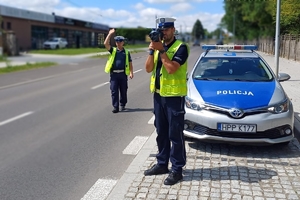 dwóch policjantów stoi przy oznakowanym radiowozie, jeden z policjantów wykonuje pomiar prędkości