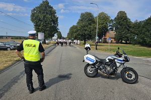 Zdjęcie przedstawia policjanta oraz policyjny motocykl na drodze.