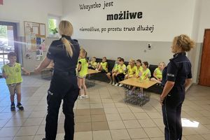 Zdjęcie przedstawia policjantki oraz dzieci w kamizelkach odblaskowych podczas prelekcji.