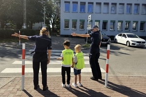 Zdjęcie przedstawia policjantki oraz dzieci w kamizelkach odblaskowych przed przejściem dla pieszych.