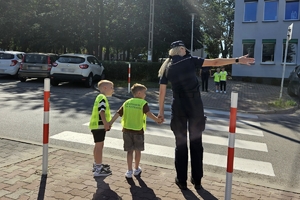 Zdjęcie przedstawia policjantkę oraz dzieci w kamizelkach odblaskowych przed przejściem dla pieszych.